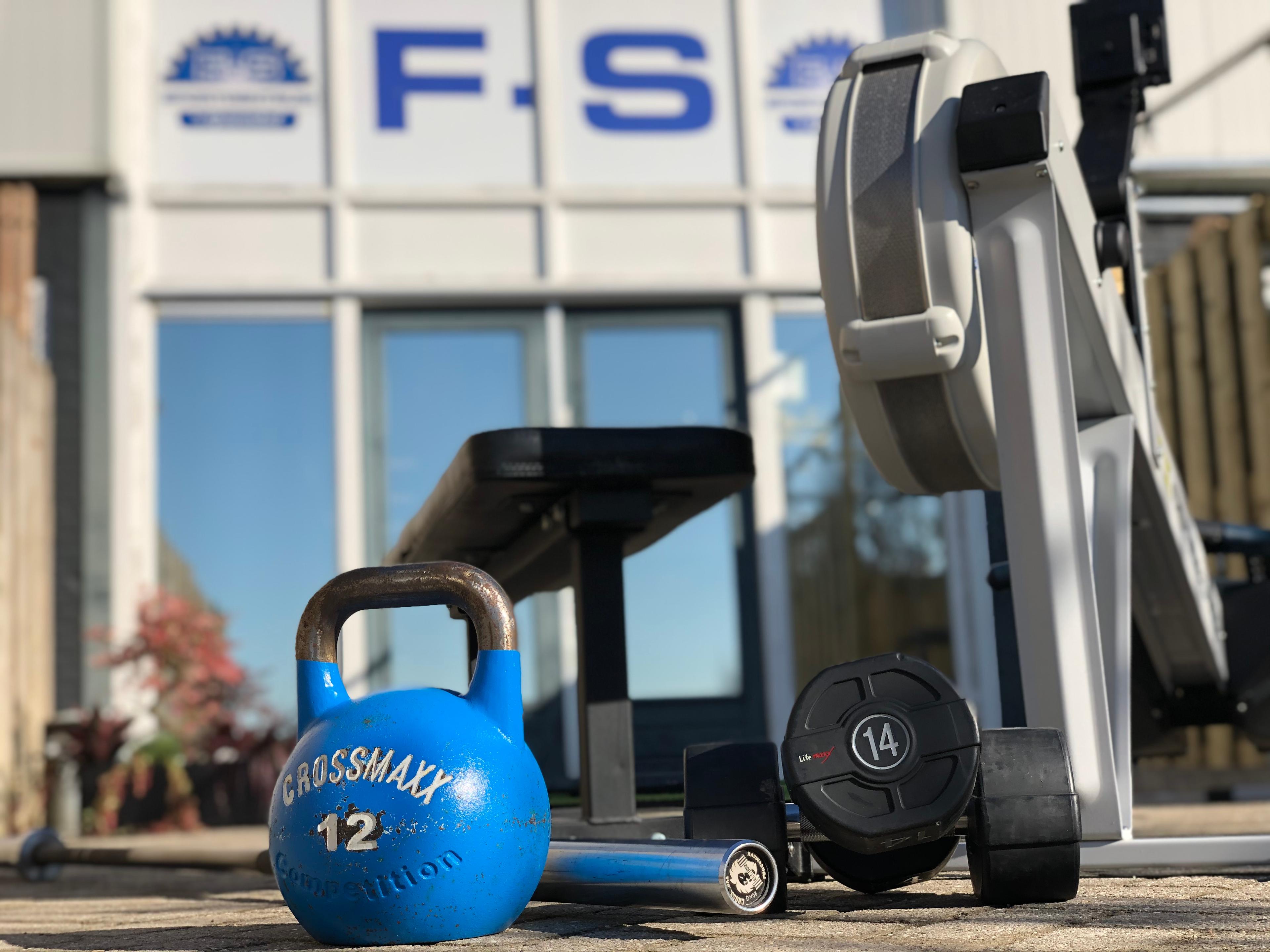 Een blauwe kettlebell, een dumbbell, en een roeimachine buiten een sportschool met het F-S Fitness logo op de achtergrond.