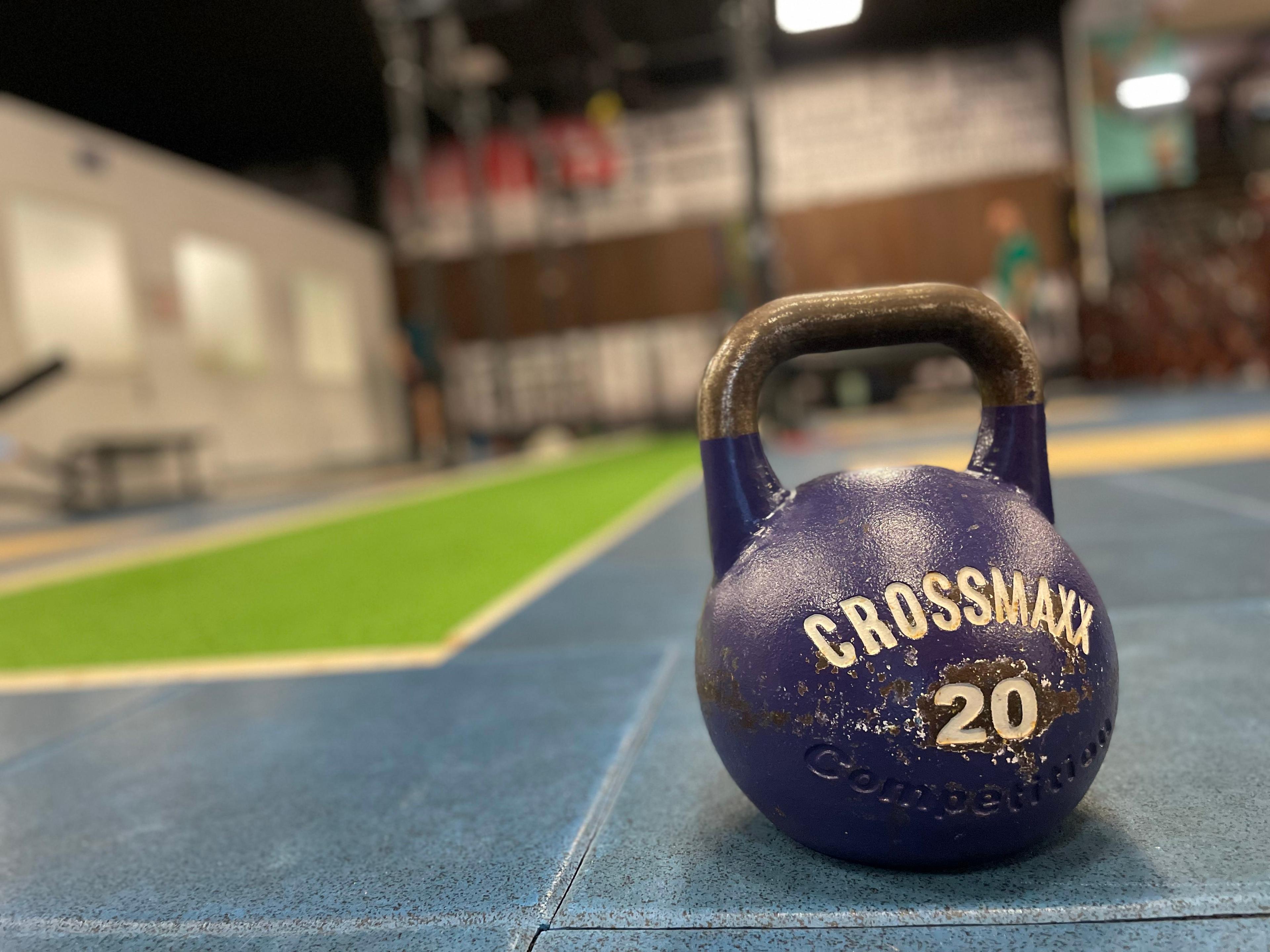 Een blauwe 20 kg Crossmaxx kettlebell staat centraal in een CrossFit-ruimte, met op de achtergrond een gedeelte van het veld en krachttrainingsapparatuur.