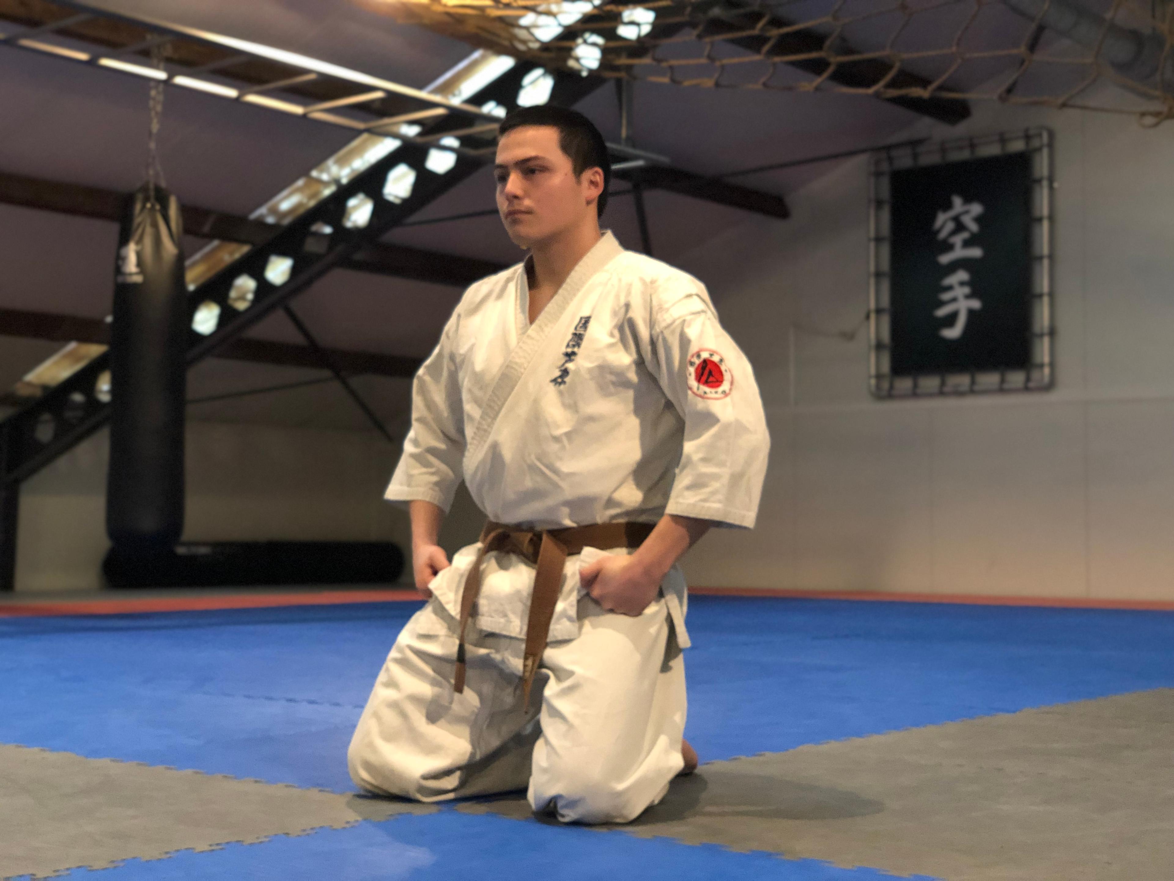 Een man in een witte karate-gi knielt op de mat in een dojo, klaar om te trainen.