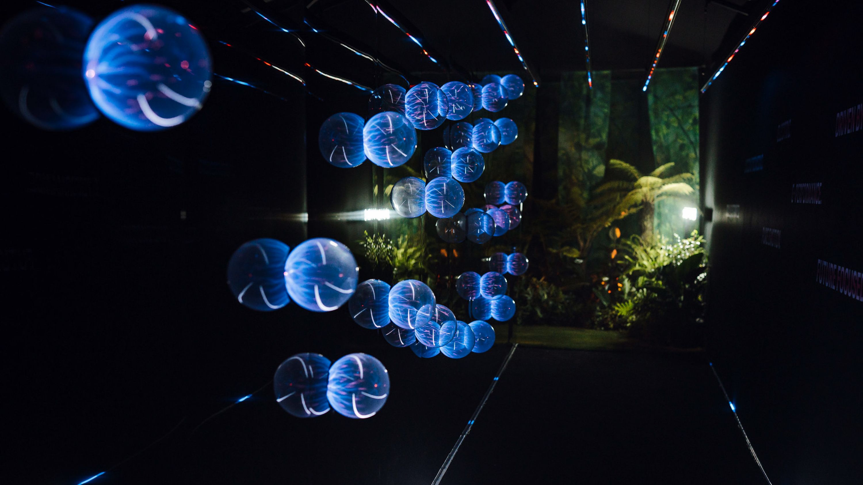 A dark hallway illuminated by blue spherical bulbs scattered on one wall, with a forest-like room revealed at the end of the hallway.