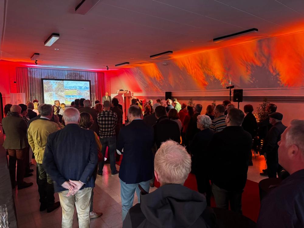 Guests standing in a red-lit hall facing a speaker at a lectern beside a projection screen