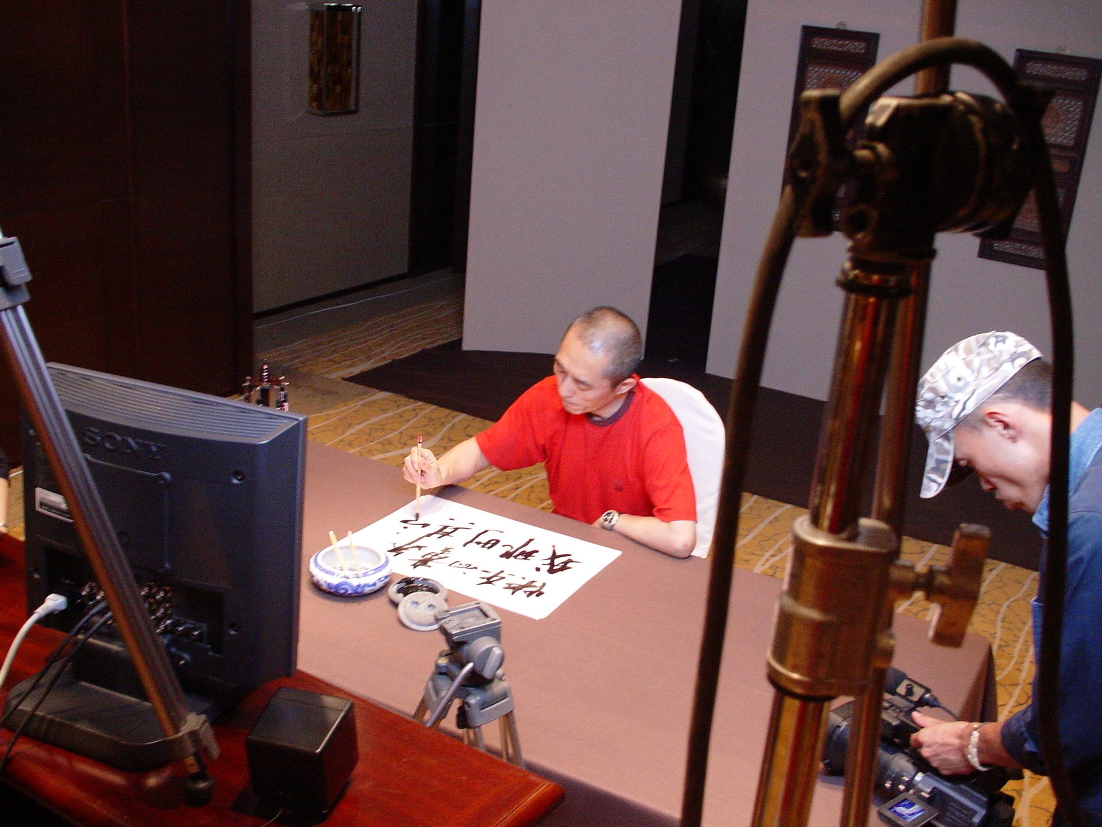High view of a man in red brushing Chinese characters on paper, filmed by a camera operator beside a Sony monitor