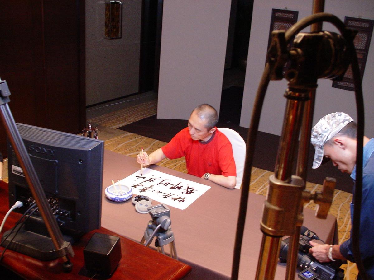 High view of a man in red brushing Chinese characters on paper, filmed by a camera operator beside a Sony monitor