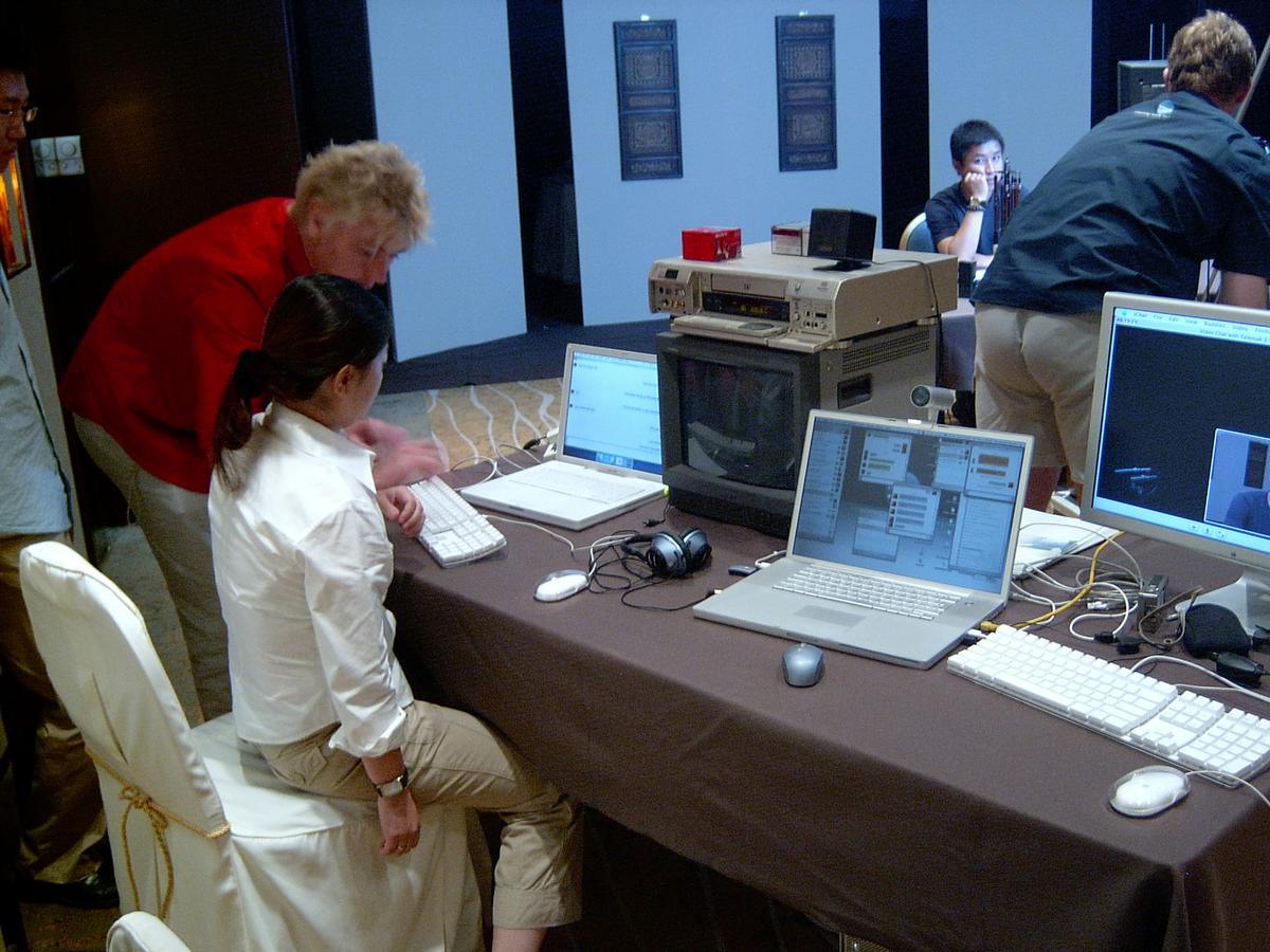 Crew at a table of laptops, a CRT monitor and a video deck while setting up the live stream in a hotel room