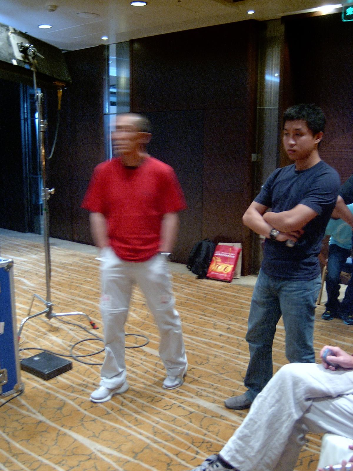 Man in a red T-shirt and a young man with folded arms standing on a hotel set beside a light stand