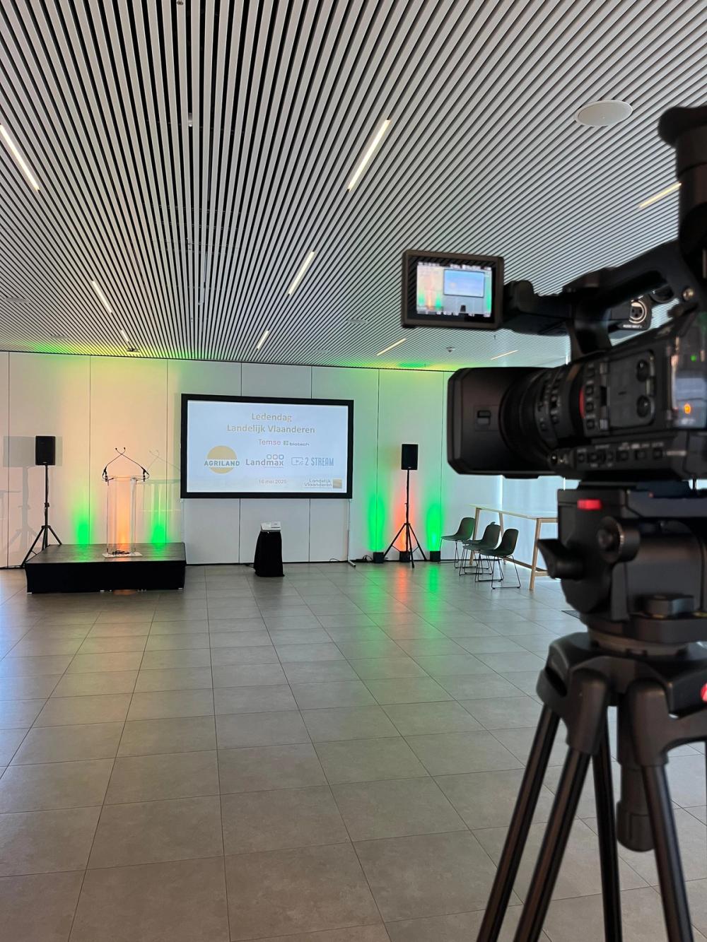 Camera on a tripod facing an empty stage with lectern and screen for the Landelijk Vlaanderen member day