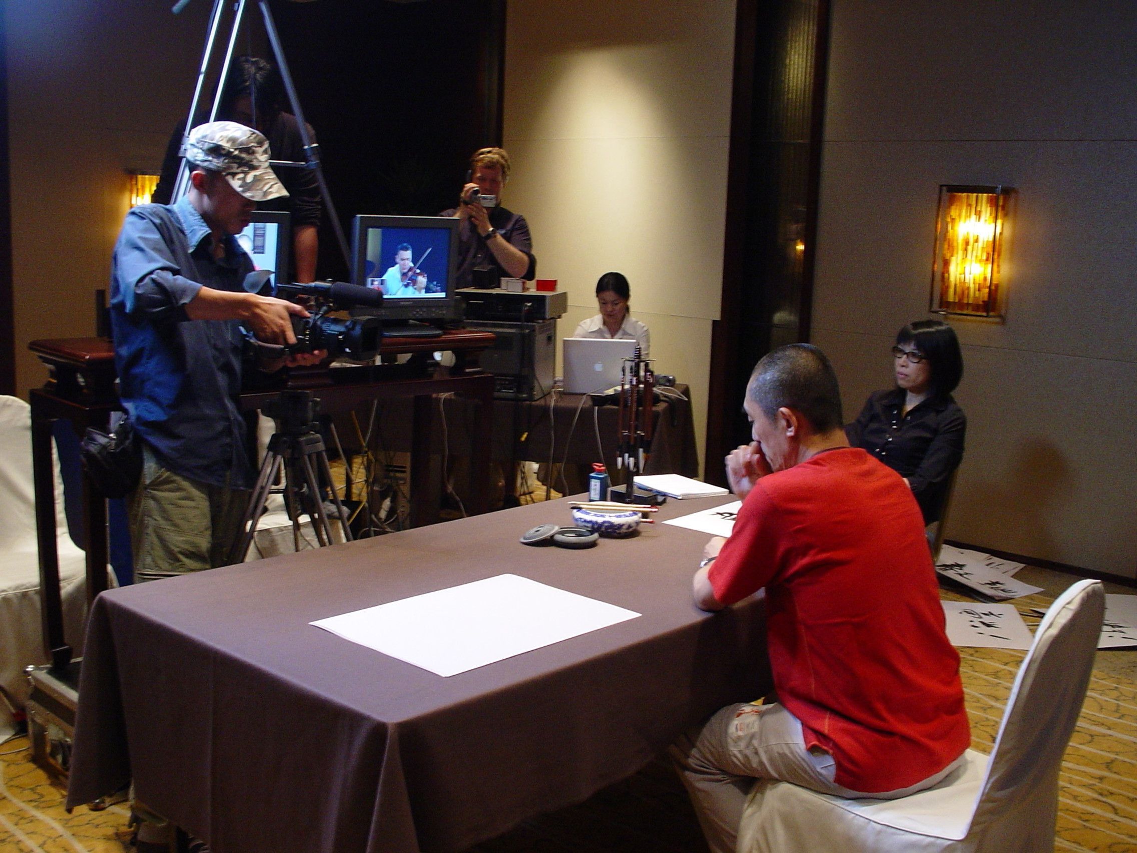 Camera operator filming a man in red at a calligraphy table, with a monitor and laptop crew behind