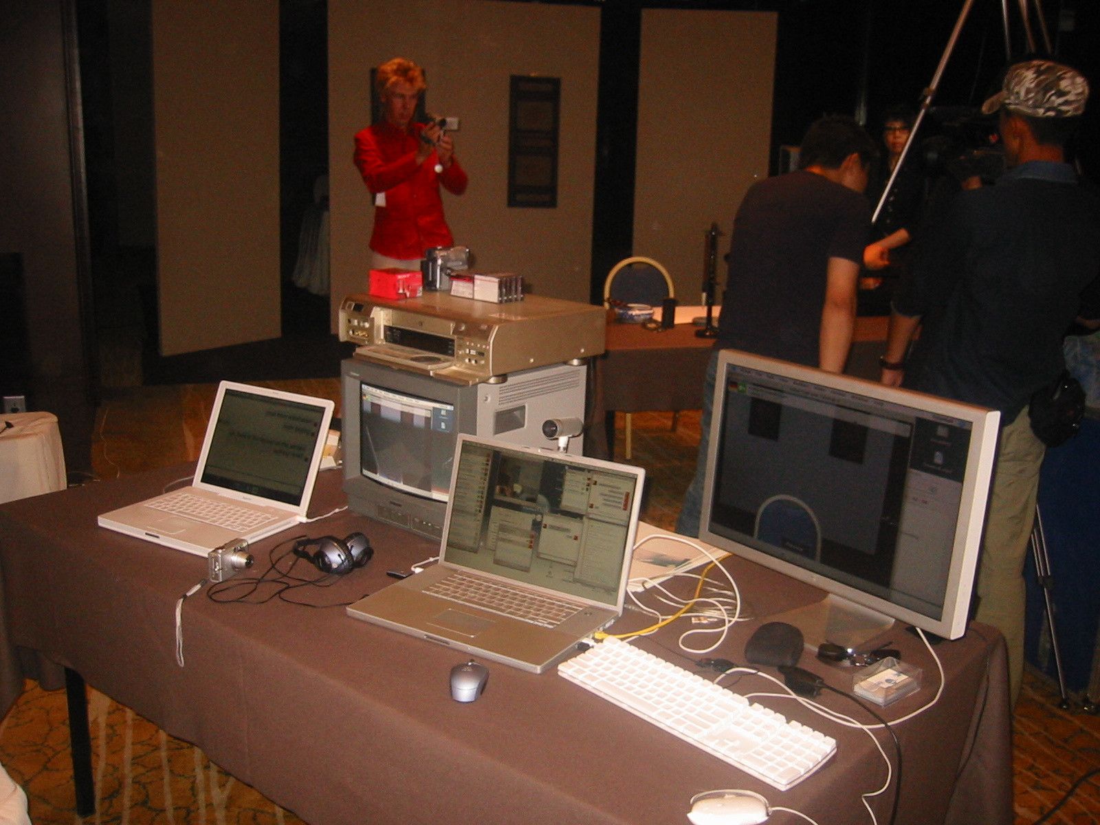 Streaming desk with laptops, a CRT monitor, a video deck and a large display, with camera operators behind