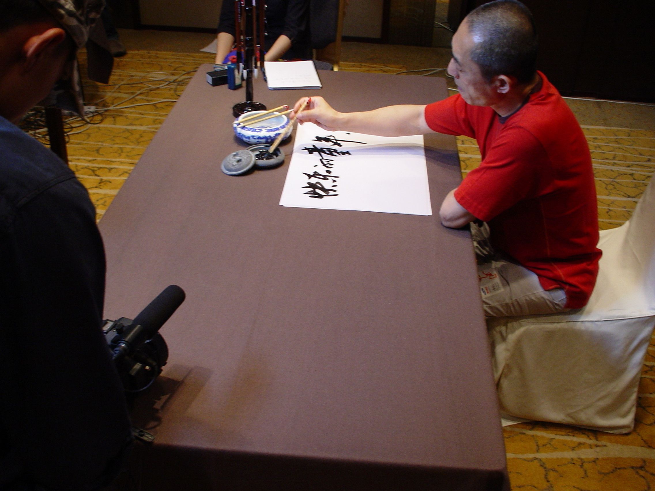 Man in a red T-shirt writing Chinese calligraphy while a camera with a shotgun microphone films across the table