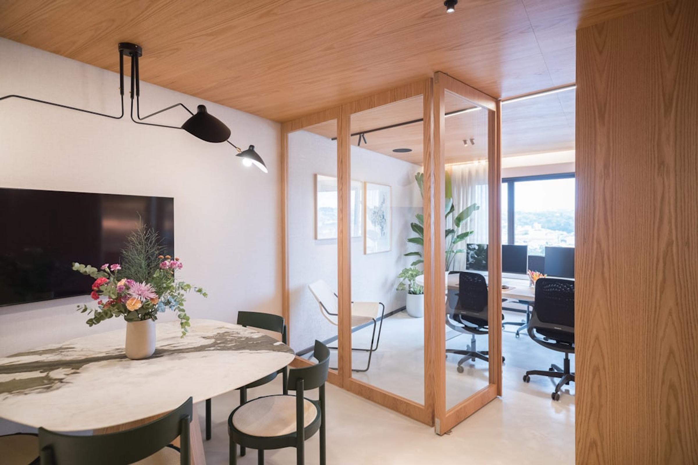 Office meeting area with a marble table and wall screen next to a glass-walled room with desks and monitors