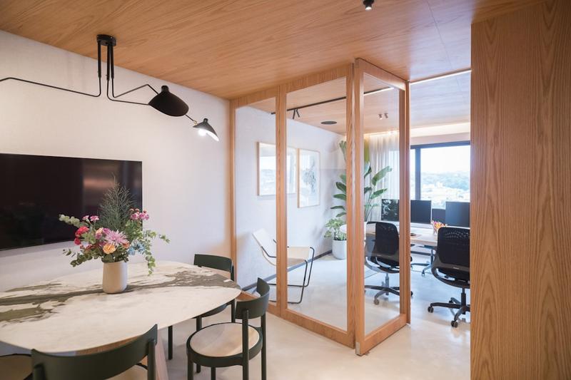 Office meeting area with a marble table and wall screen next to a glass-walled room with desks and monitors
