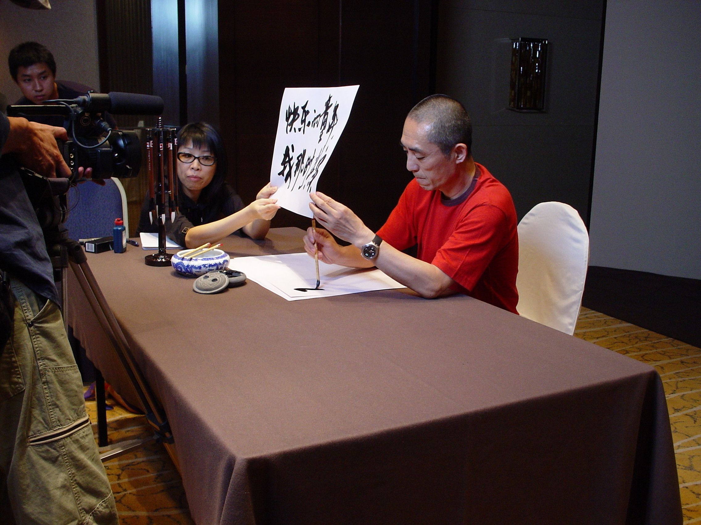 Man in a red T-shirt holding up a sheet of Chinese calligraphy for the camera, a woman seated beside him
