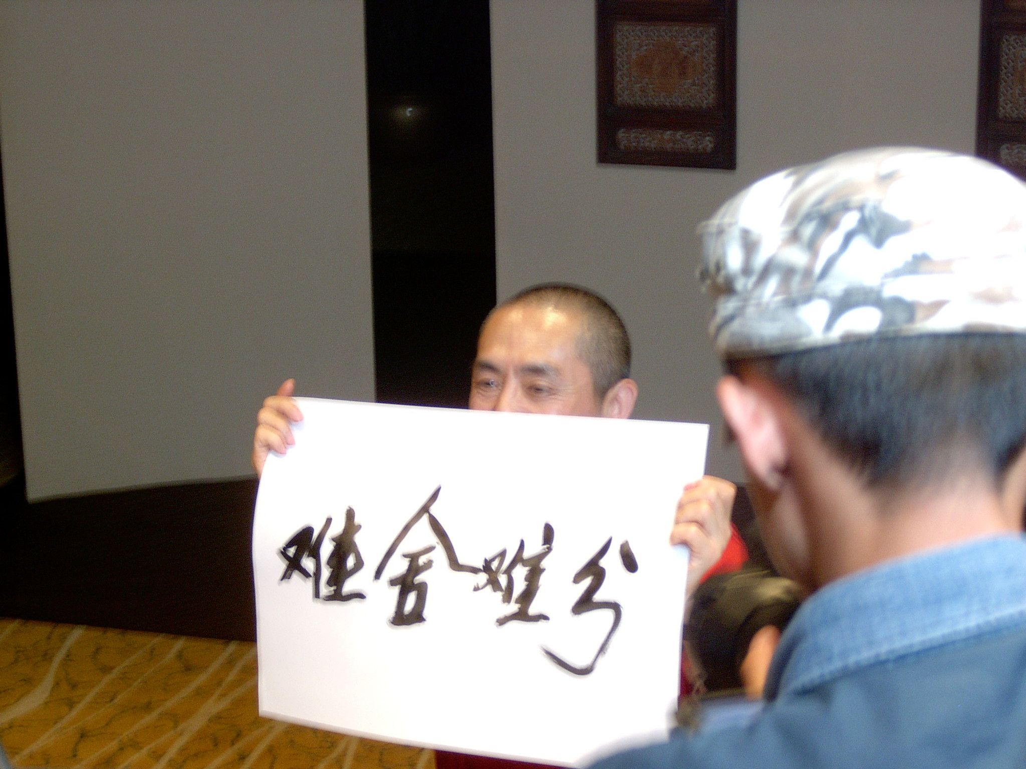 Man holding up a sheet of Chinese calligraphy in front of a crew member wearing a patterned cap