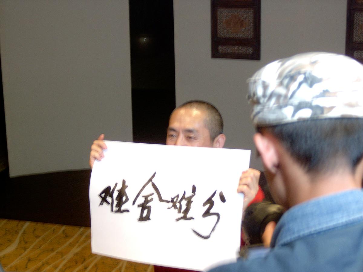 Man holding up a sheet of Chinese calligraphy in front of a crew member wearing a patterned cap