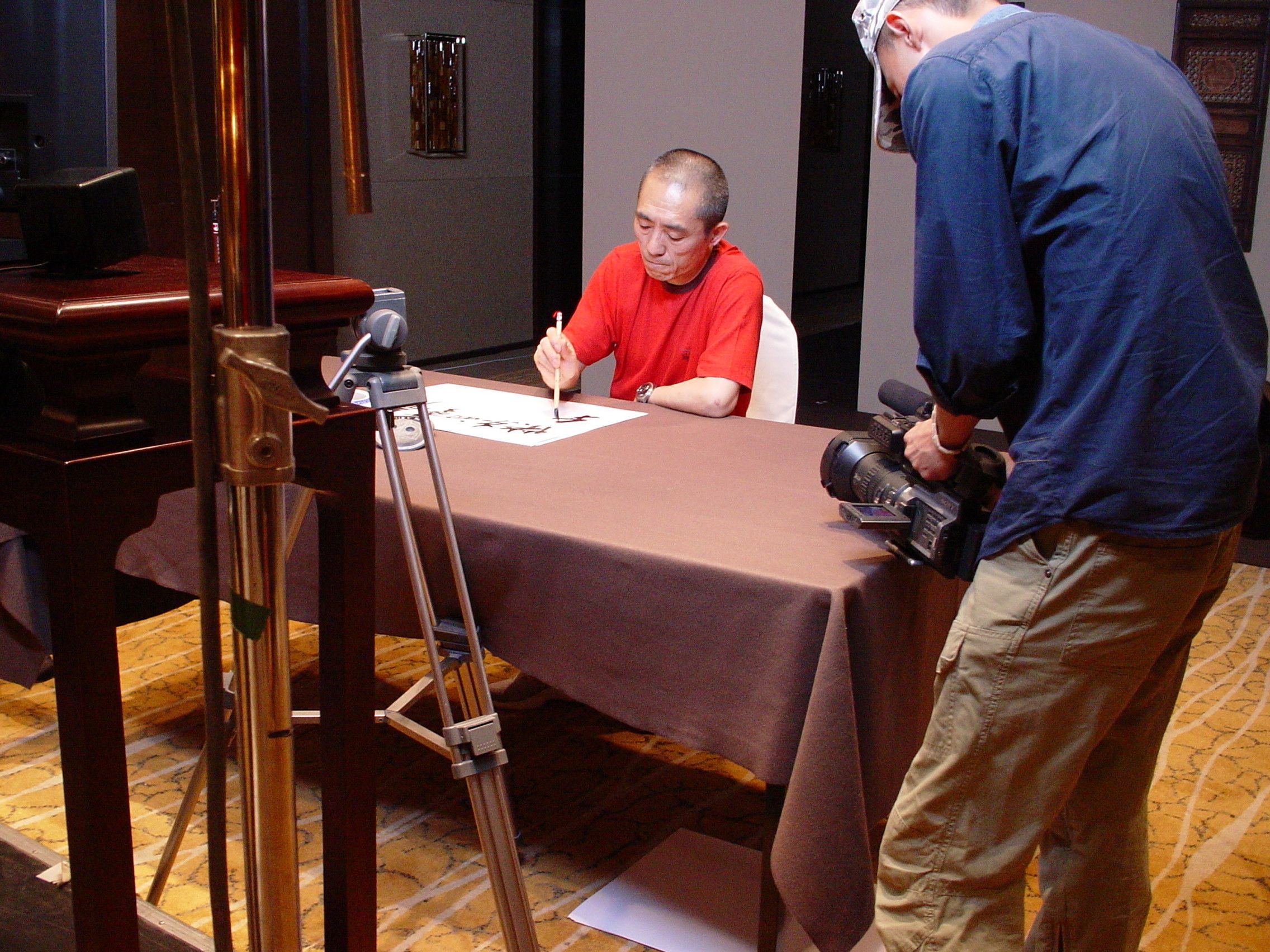 Man in a red T-shirt writing Chinese calligraphy with a brush while a camera operator films him up close