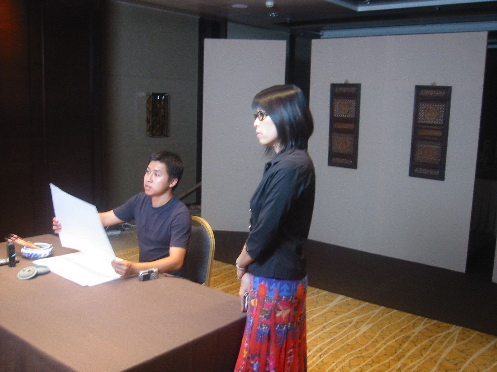 Young man at a table holding up a large white sheet while a woman in glasses stands beside him on set