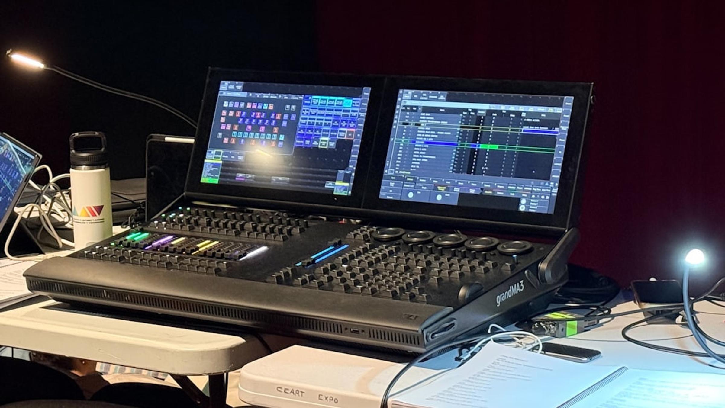 grandMA3 lighting console with two touchscreens on a production desk beside a gooseneck lamp