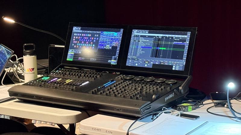 grandMA3 lighting console with two touchscreens on a production desk beside a gooseneck lamp