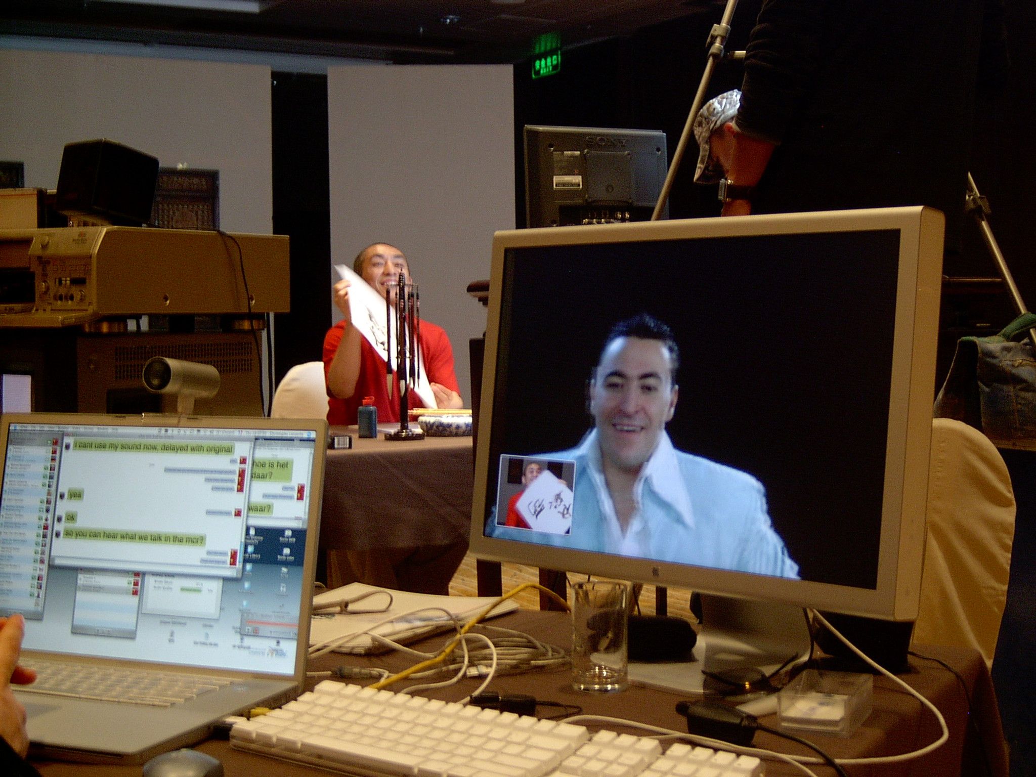 Monitor showing a smiling remote participant while a man in red holds up calligraphy in the room behind