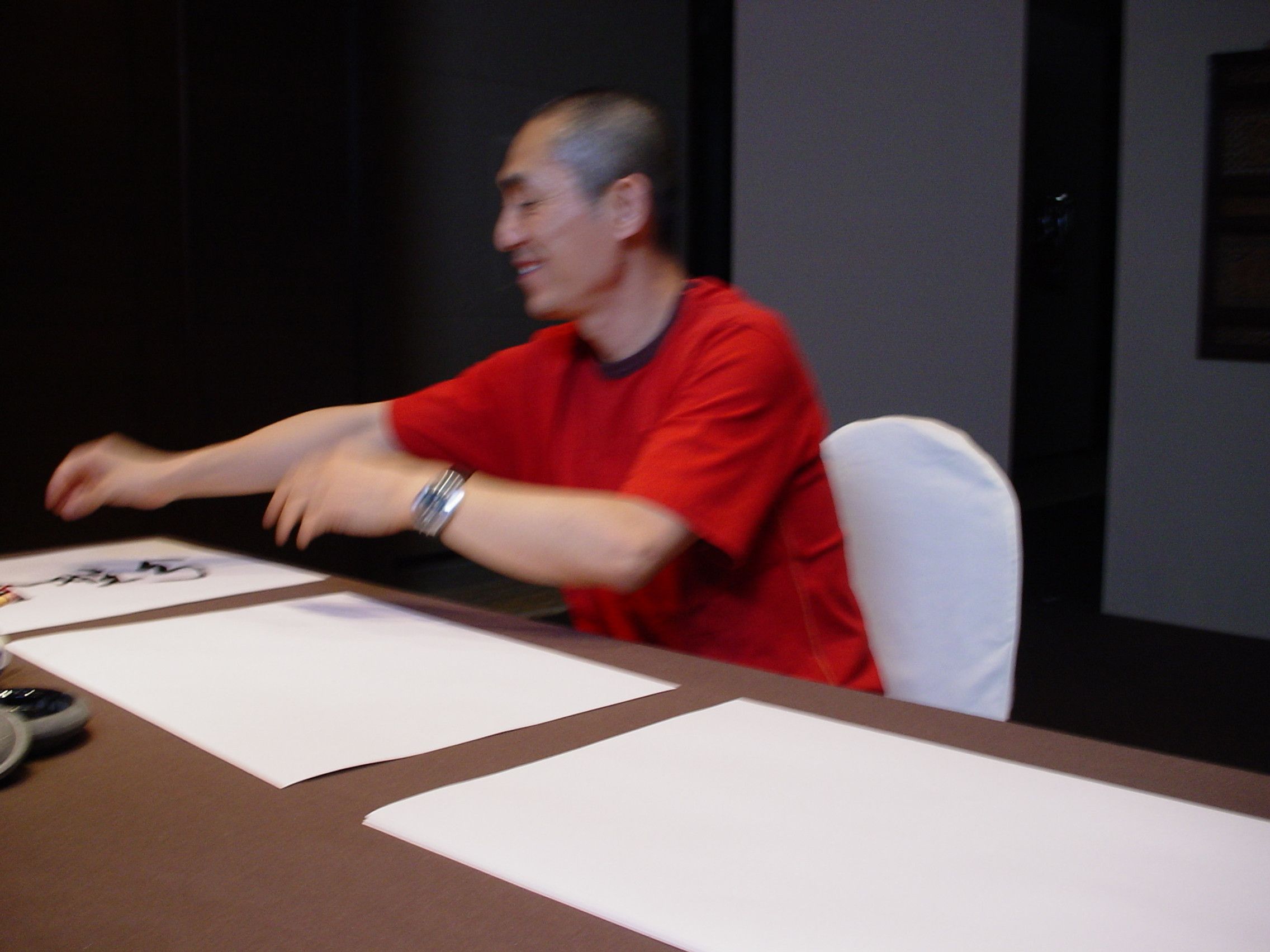 Man in a red shirt laughing and sweeping his arms across a table of blank calligraphy paper