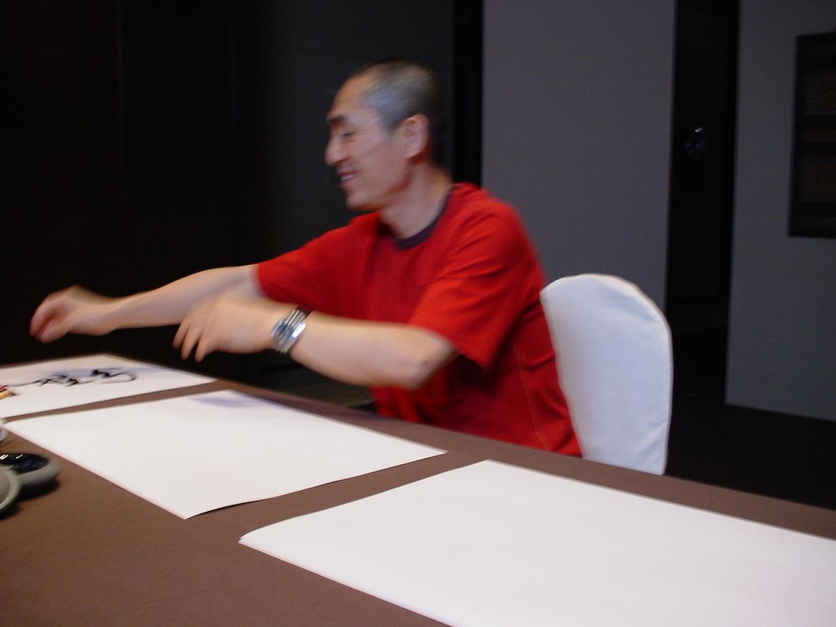 Man in a red shirt laughing and sweeping his arms across a table of blank calligraphy paper