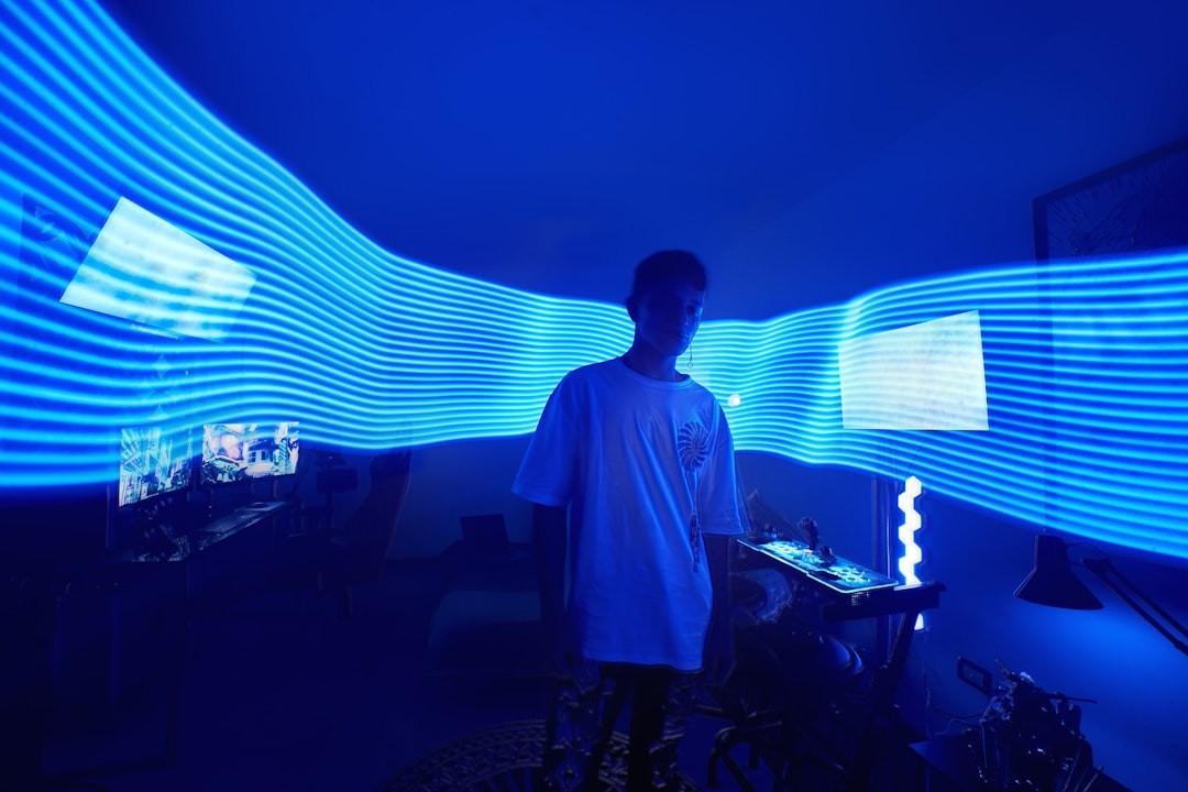 Person in a white T-shirt surrounded by blue light-painting streaks in a dark room with screens