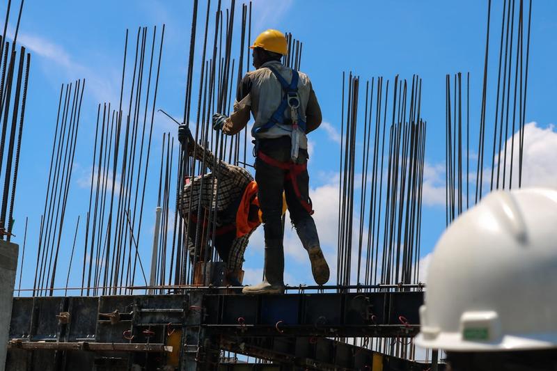 Construction workers in hard hats and safety harnesses on formwork among steel rebar against a blue sky