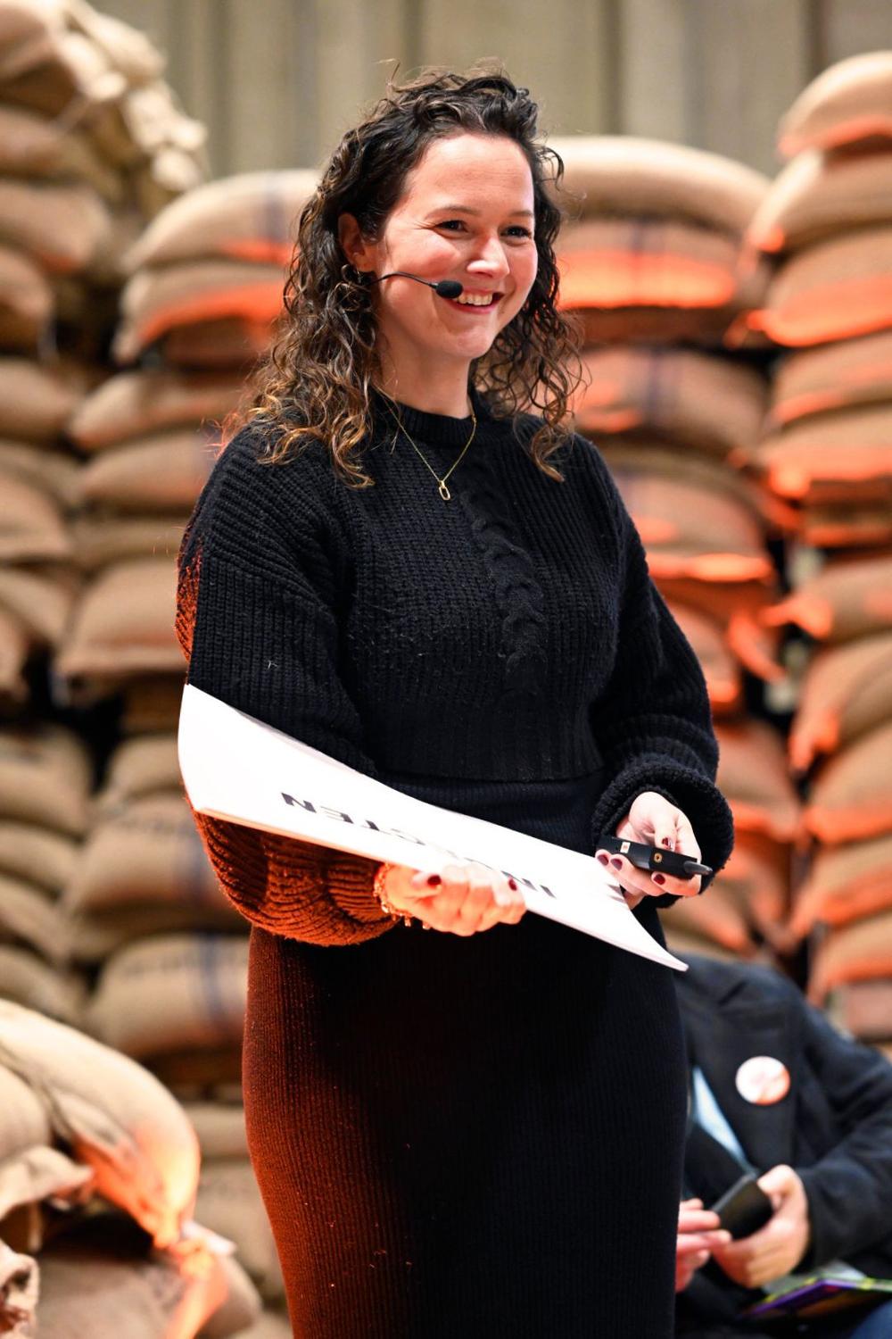 Smiling presenter with a headset microphone holding a script on a stage lined with stacked jute sacks
