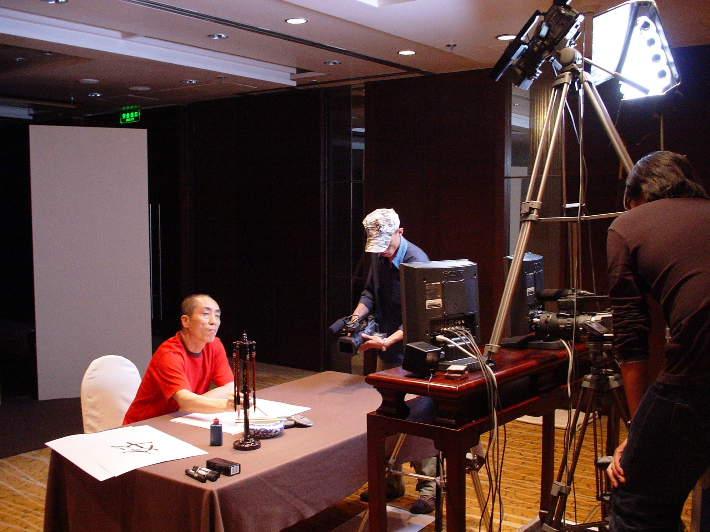 Man in a red shirt at a calligraphy table facing a camera operator, monitors and studio lights