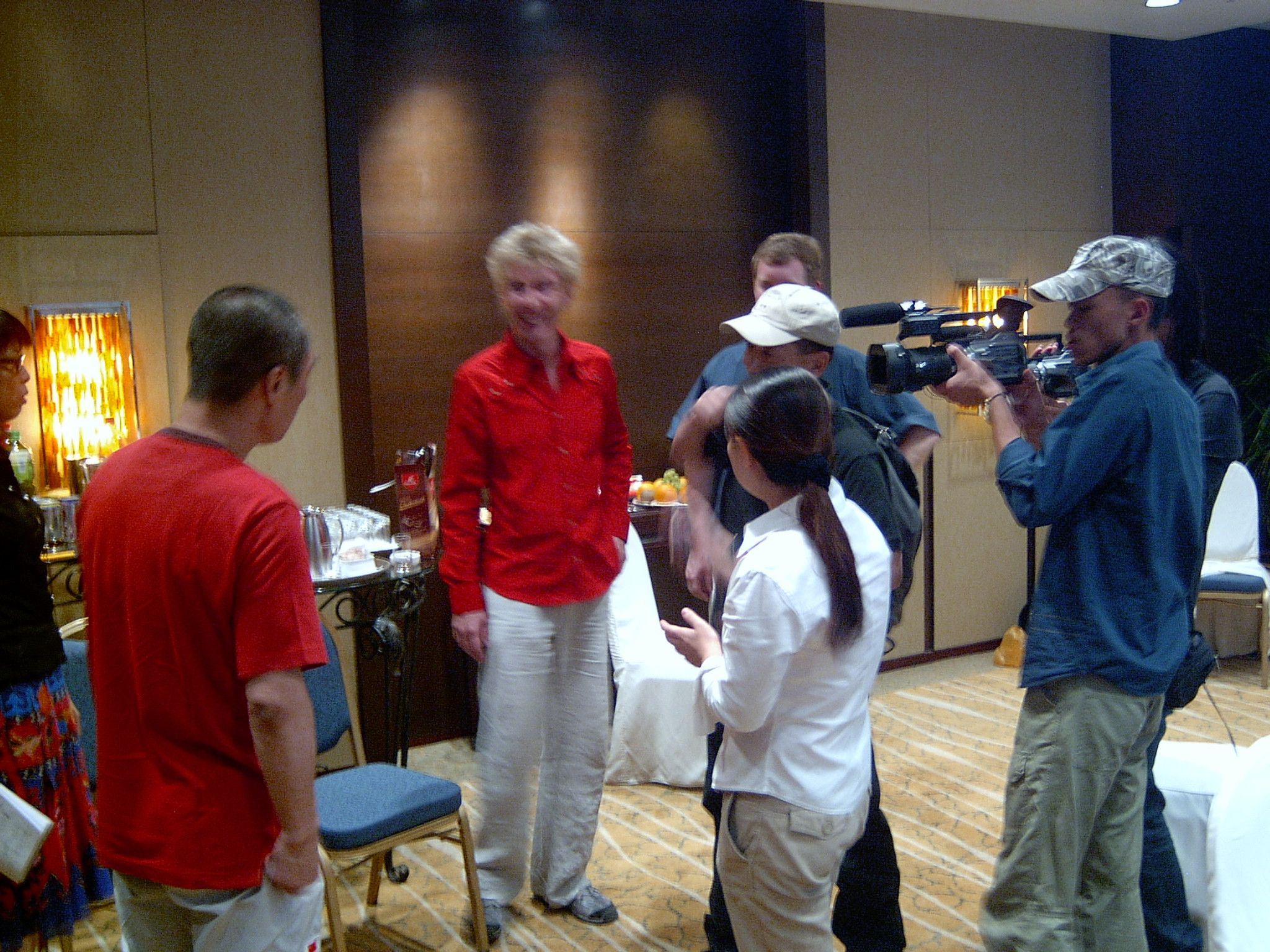 Camera operator filming a woman in a red shirt as she talks with a small group in a hotel lounge