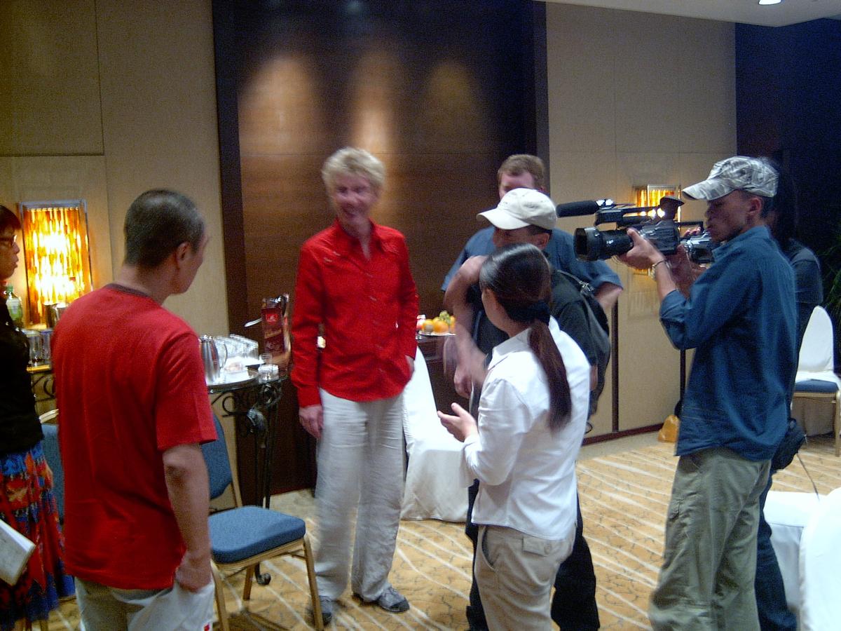 Camera operator filming a woman in a red shirt as she talks with a small group in a hotel lounge