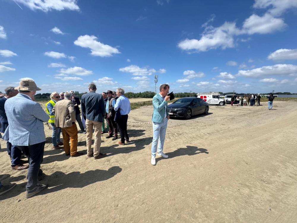 Guests gathered on a sandy open site under a blue sky, with cars and a white van in the background