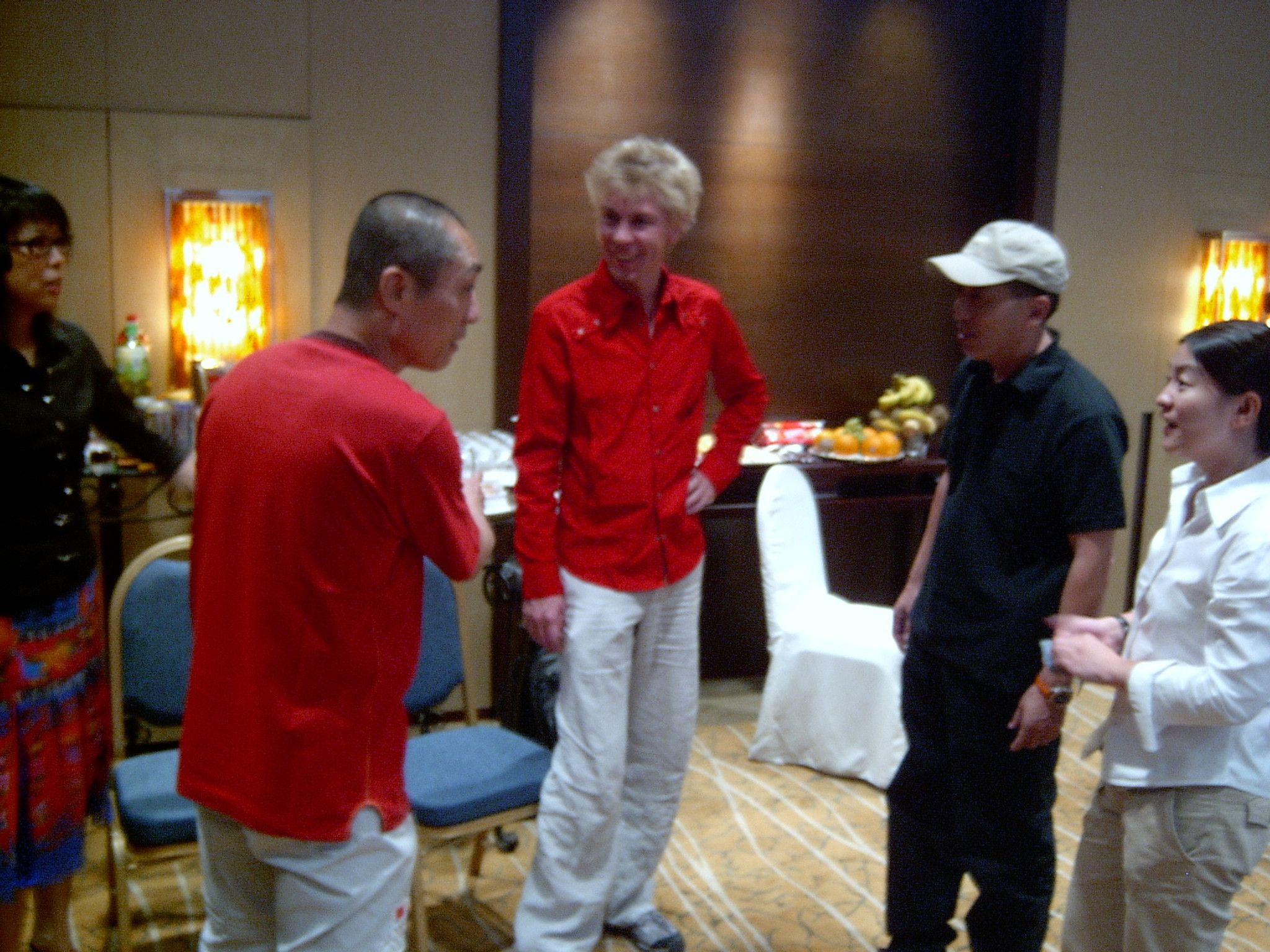 Five people talking in a hotel function room, among them a man in a red shirt and a man in a white cap