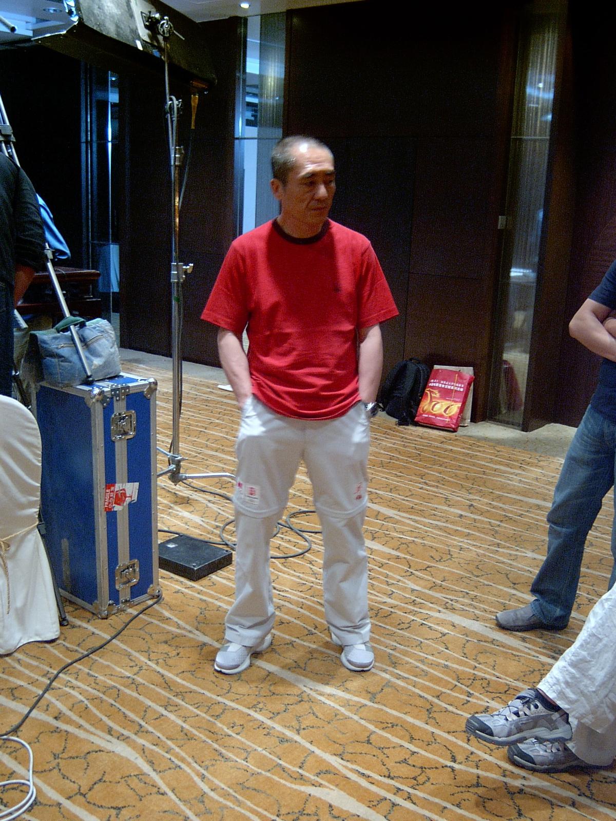 Man in a red T-shirt and white trousers standing among flight cases and light stands on set