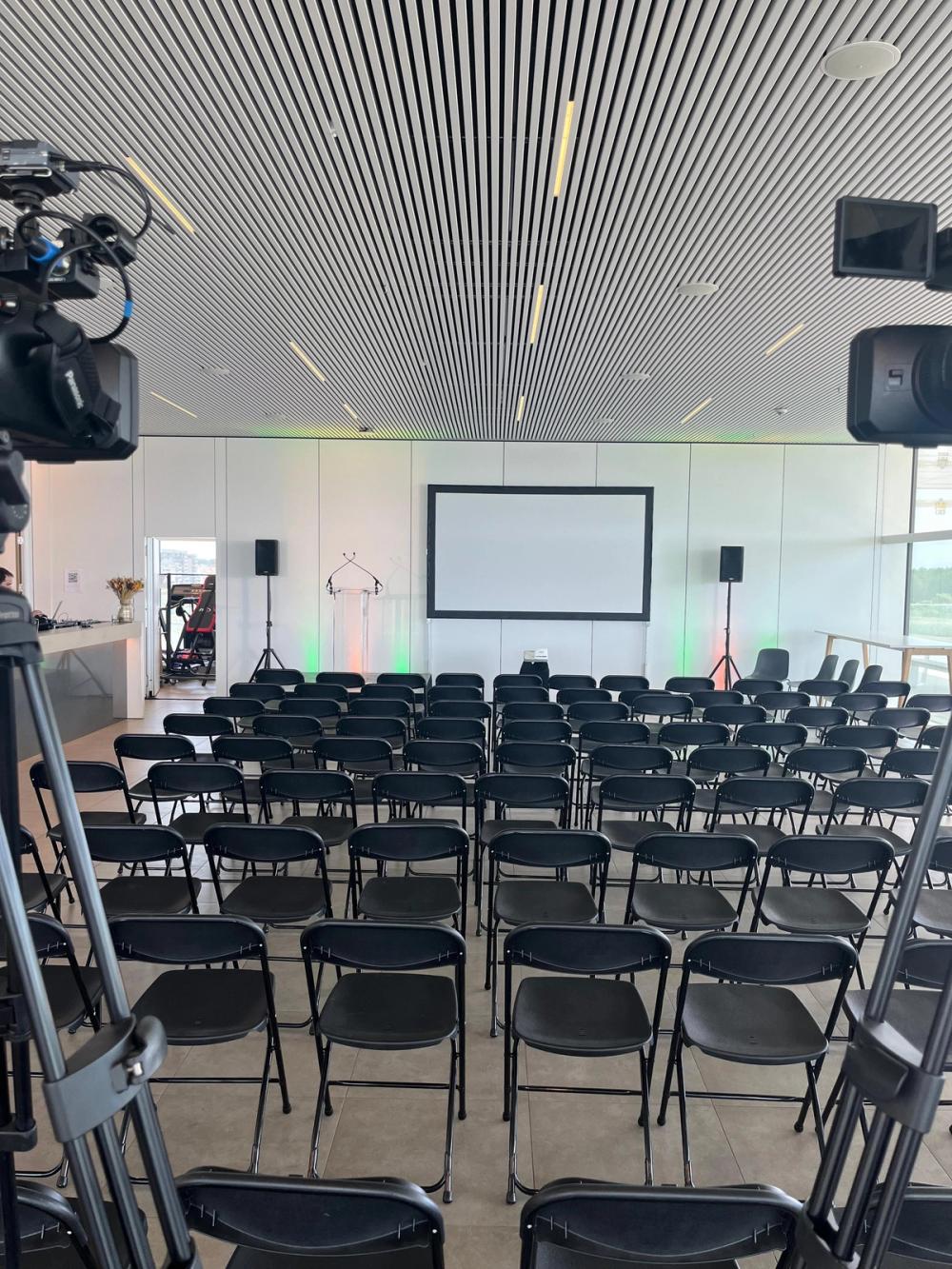 Rows of empty chairs facing a projection screen and lectern, seen between two cameras on tripods before the event