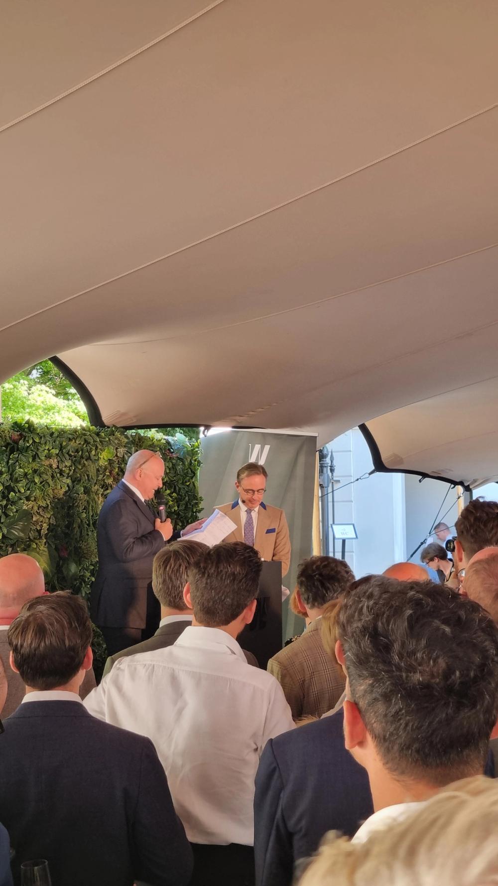 Two speakers at a lectern under a tent canopy at De Warande, addressing a standing audience