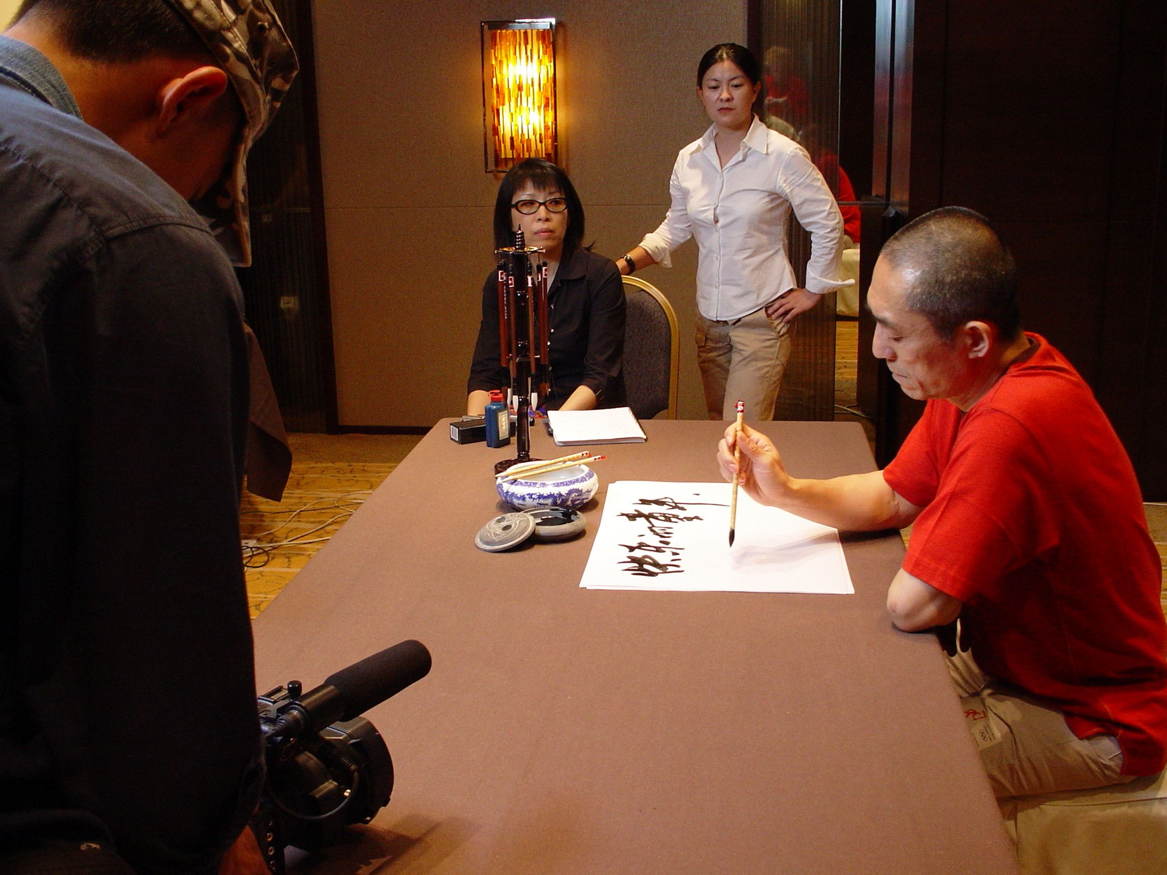 Camera operator filming a man in red painting calligraphy as two women watch from the end of the table