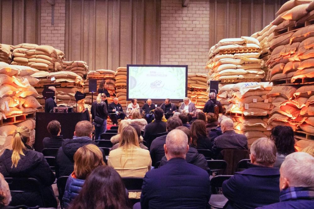 Audience facing a panel on a small stage in a warehouse lined with stacked sacks at a Sustainable Stories event
