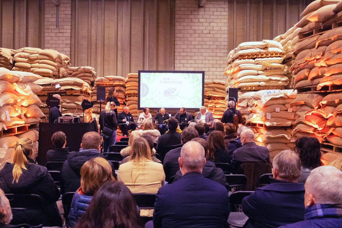 Audience watching a panel in a warehouse stacked with jute sacks, the screen showing Spin wordt Klimaatheldin