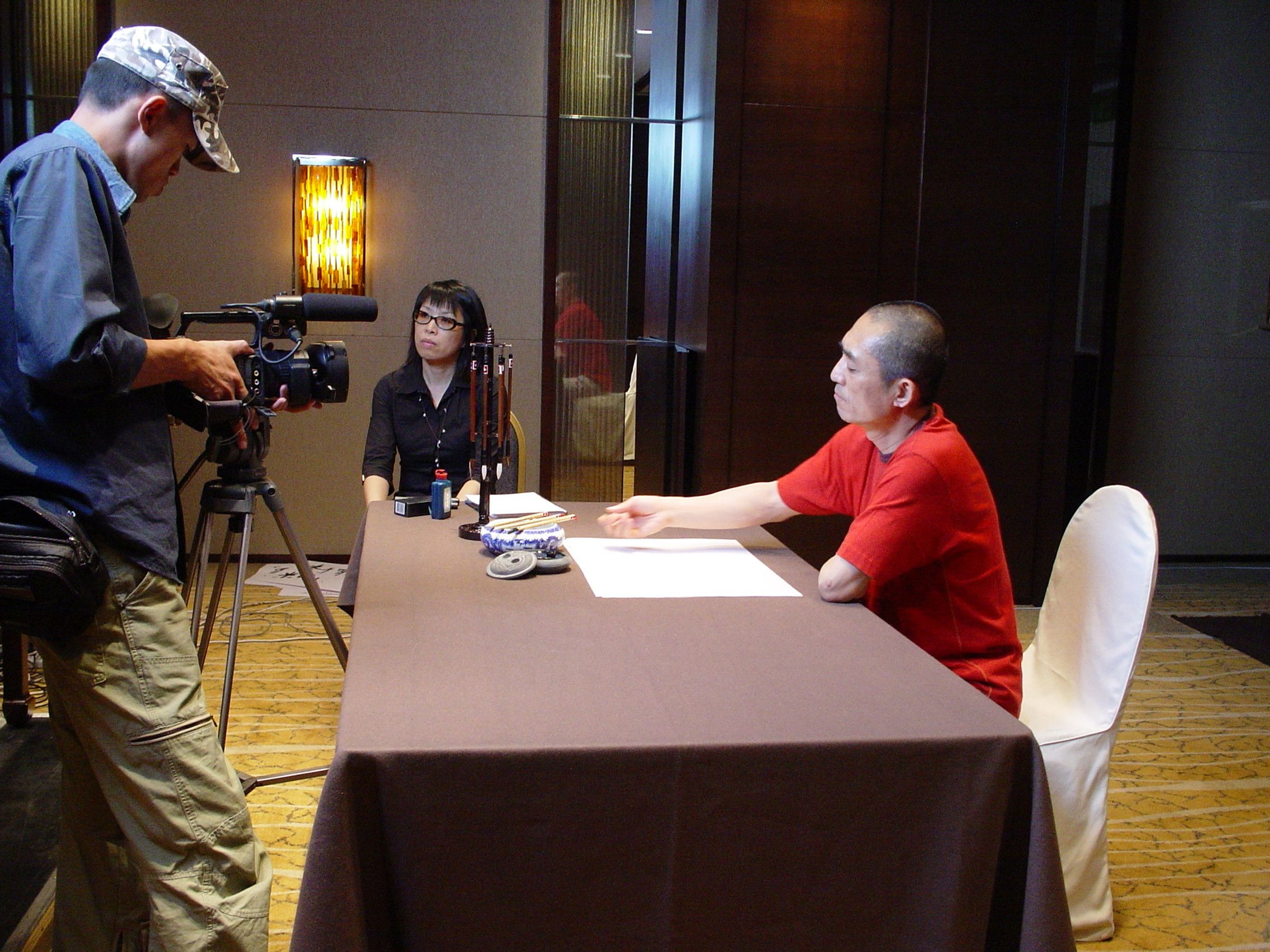 Camera operator filming a man in a red shirt at a table, as a woman in glasses watches from the end