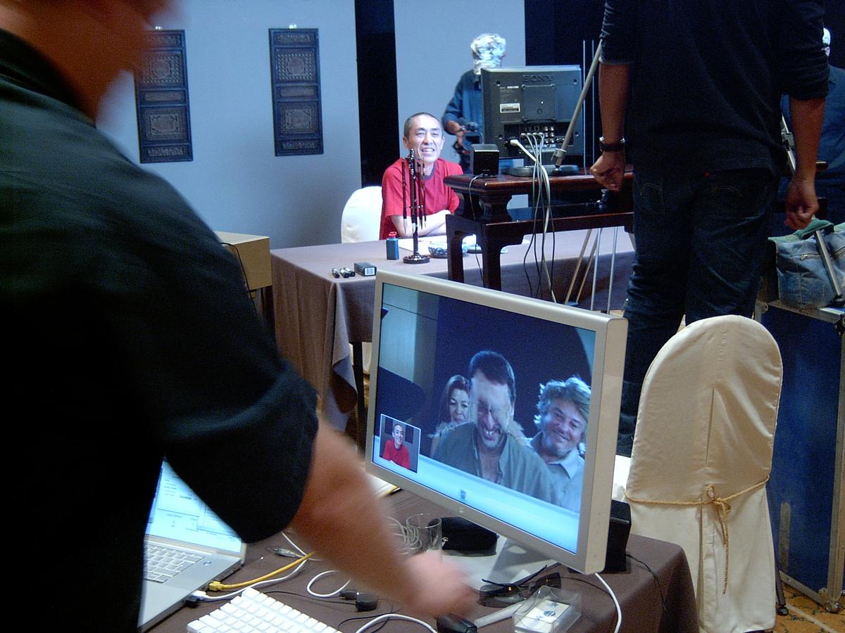 Video call on a monitor showing three people laughing, with the man in red at the calligraphy table behind