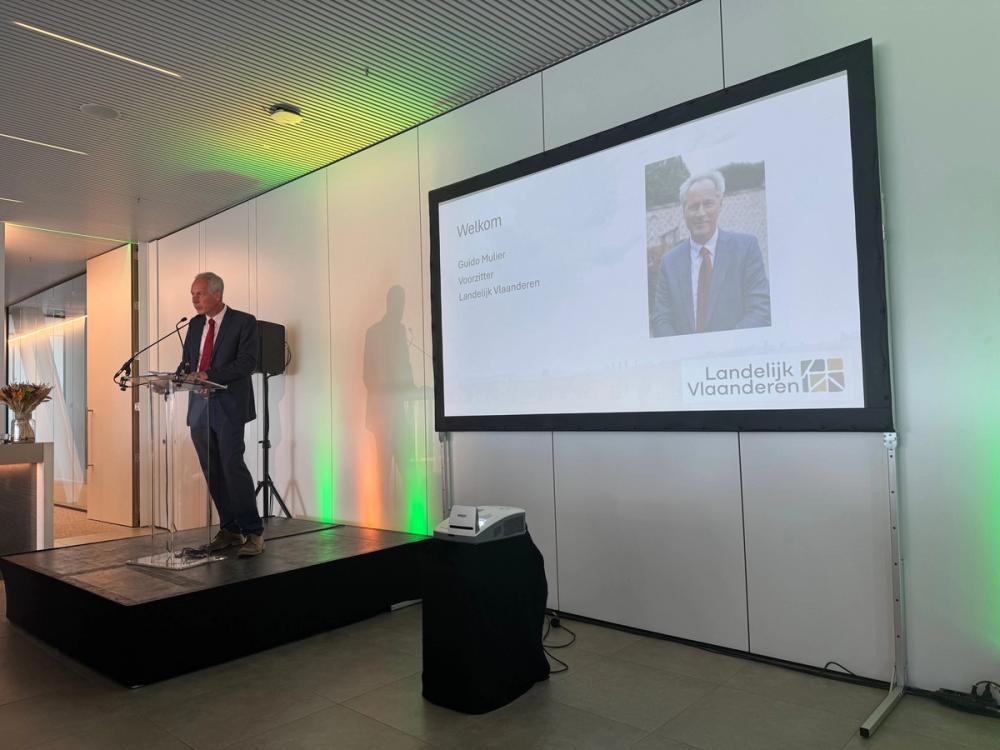 Guido Mulier, chair of Landelijk Vlaanderen, speaking at a lectern beside a welcome slide with his photo