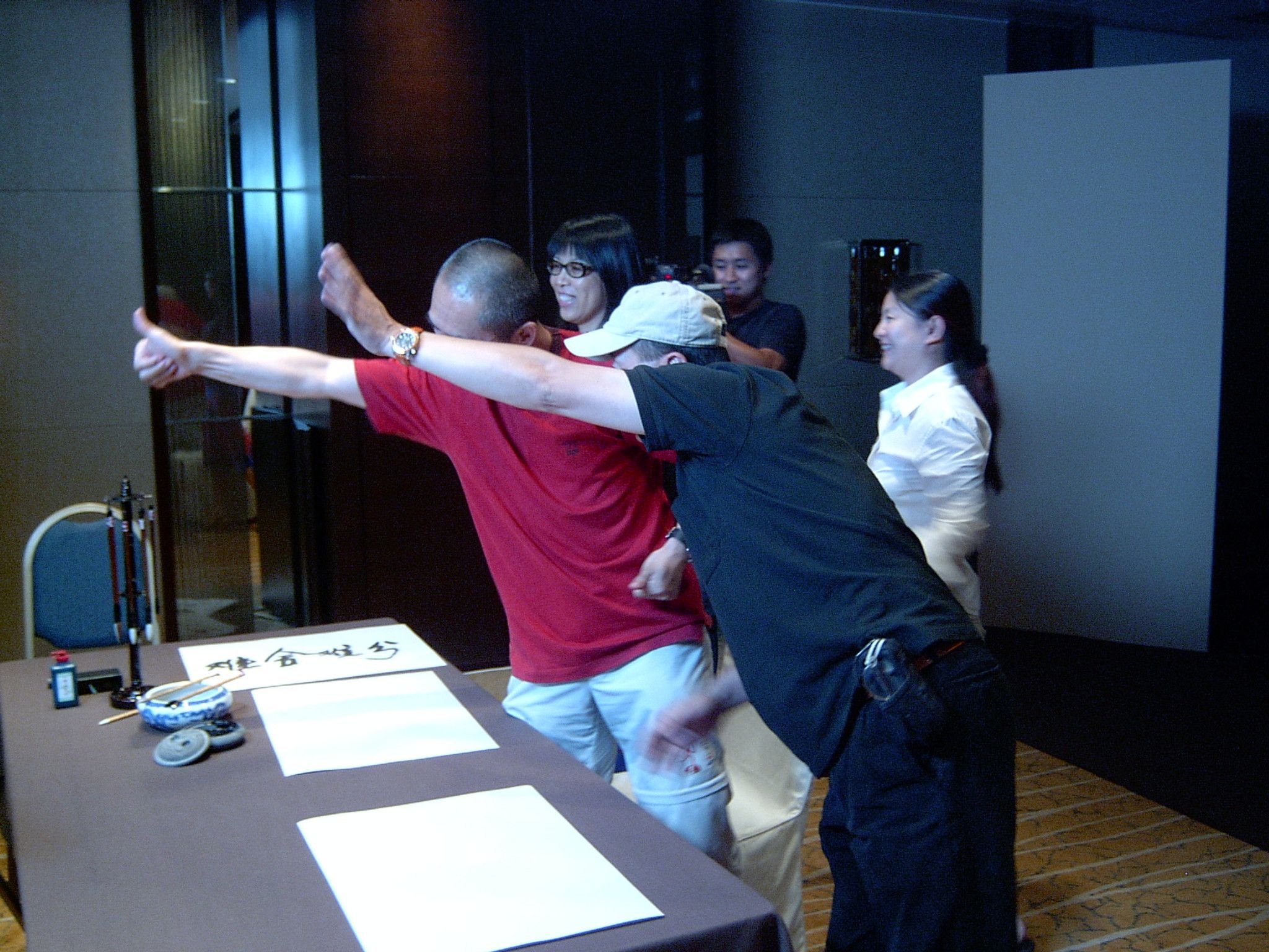 Man in a red shirt and a man in a cap stretching out their arms beside a calligraphy table, laughing