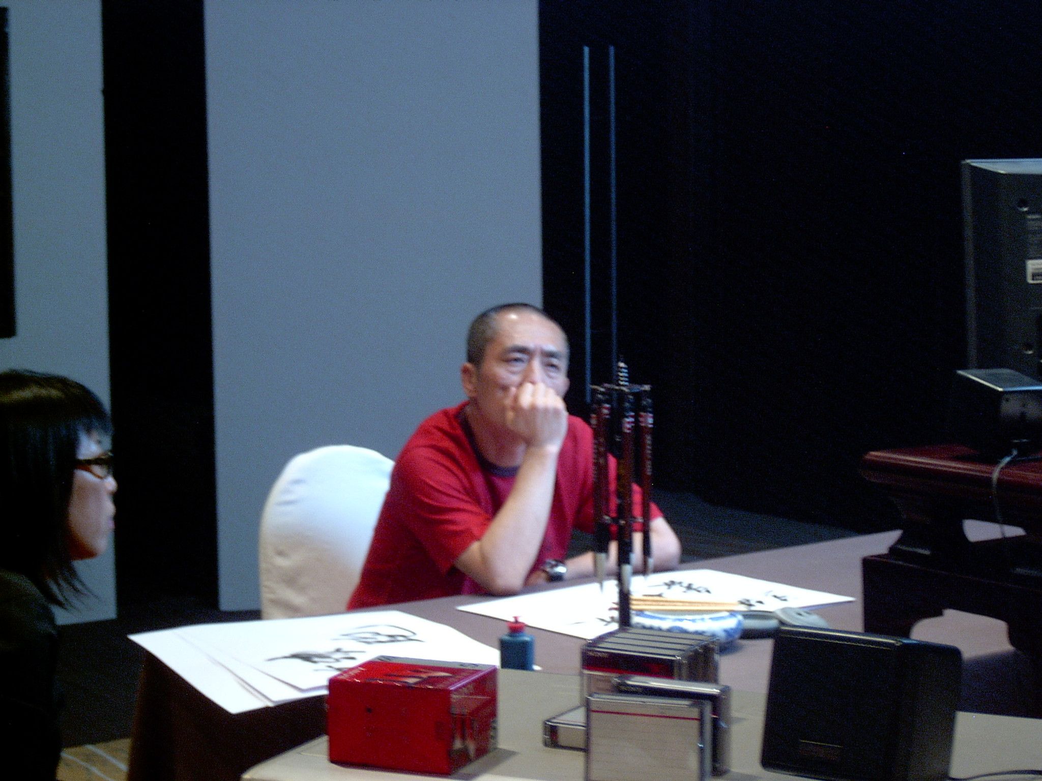Man in a red T-shirt at a table with calligraphy brushes and inked paper, watching a monitor off to the side