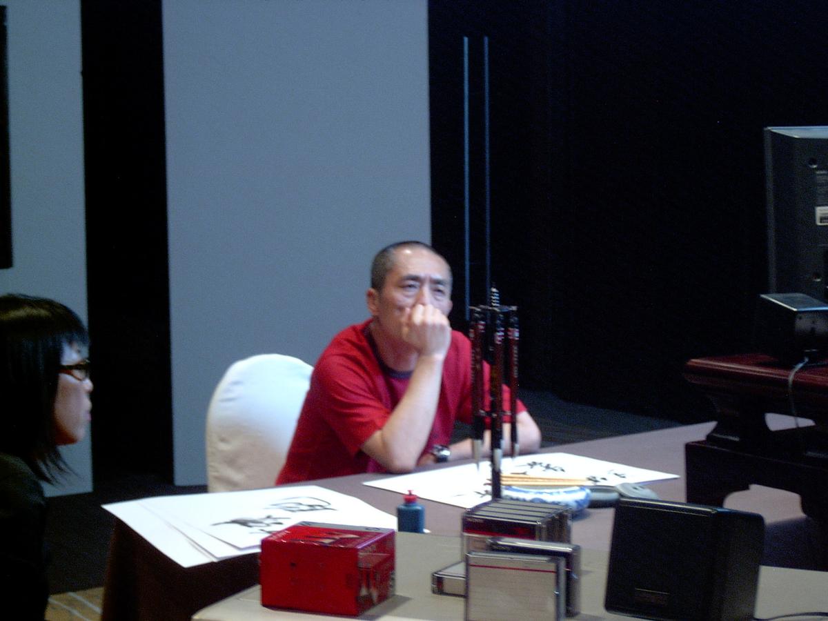 Man in a red T-shirt at a table with calligraphy brushes and inked paper, watching a monitor off to the side