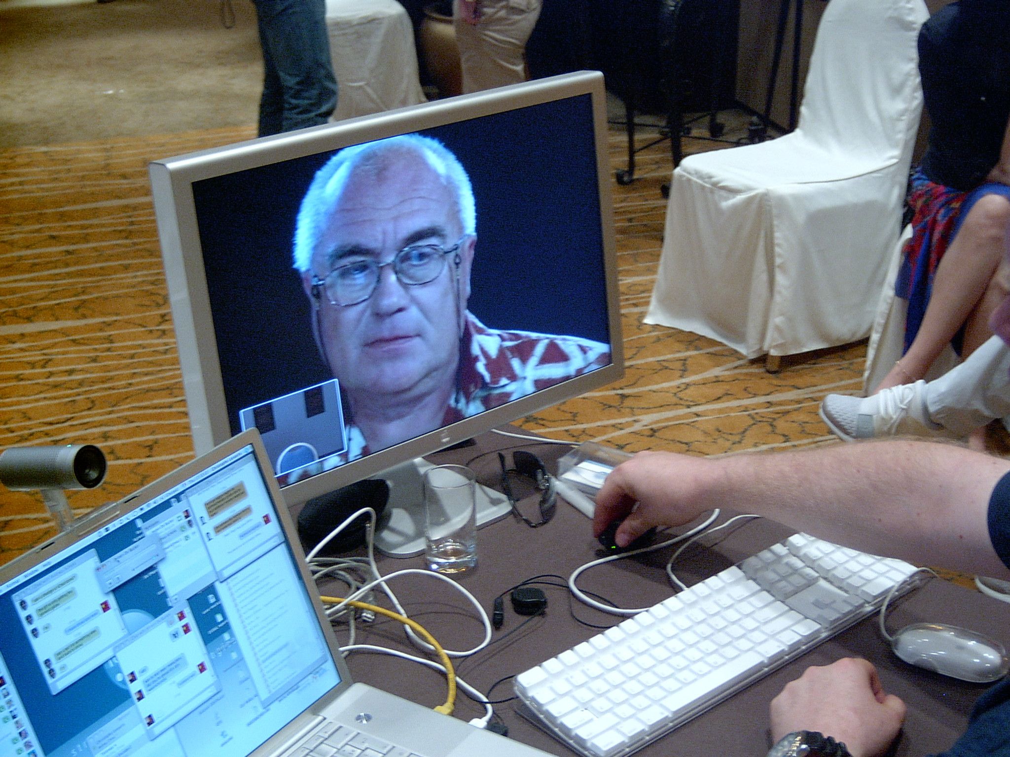 Operator's hand on a mouse beside a laptop chat window and a monitor showing a remote participant on video