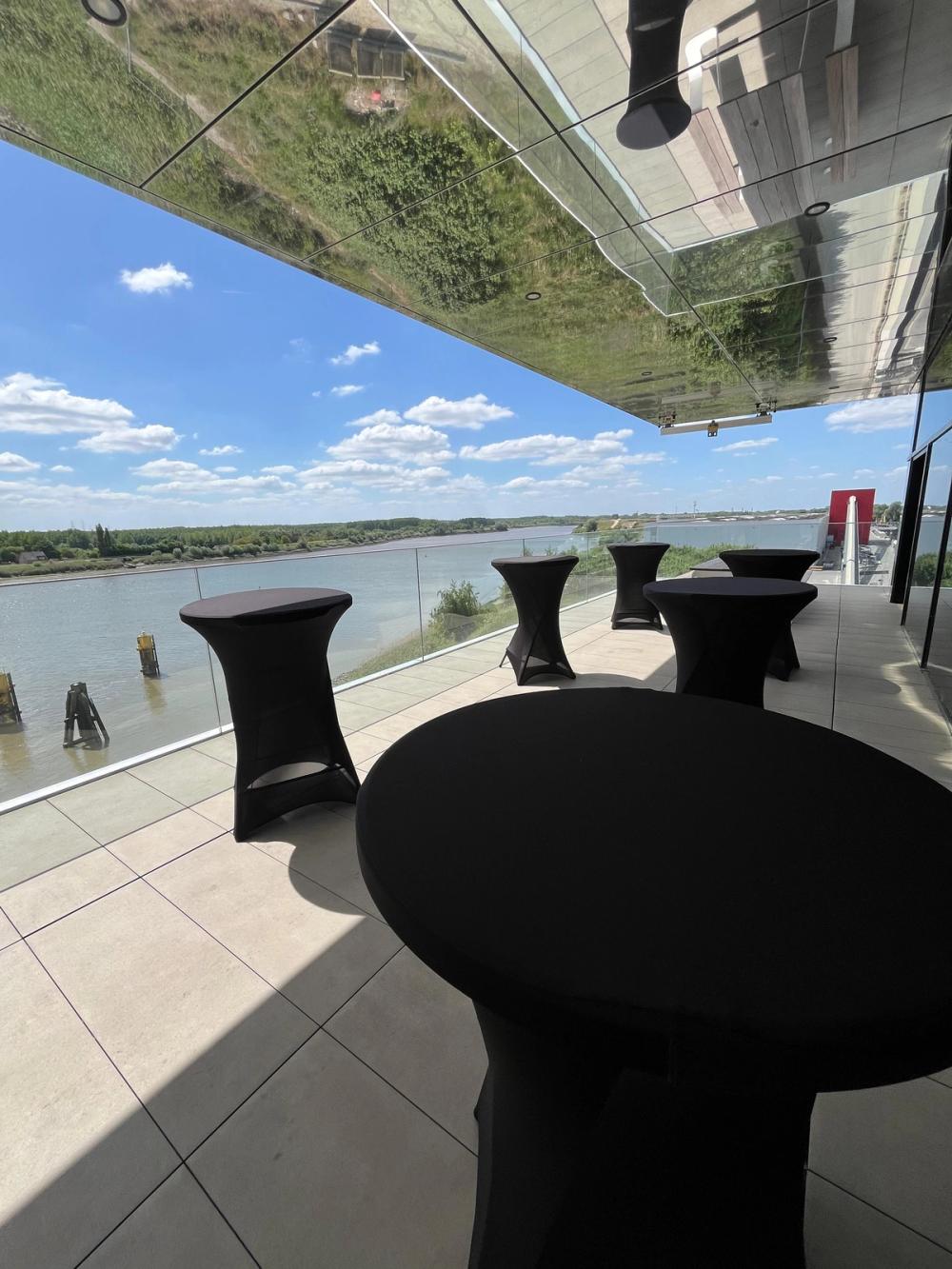 Riverside terrace with black-draped standing tables under a mirrored canopy on a sunny day