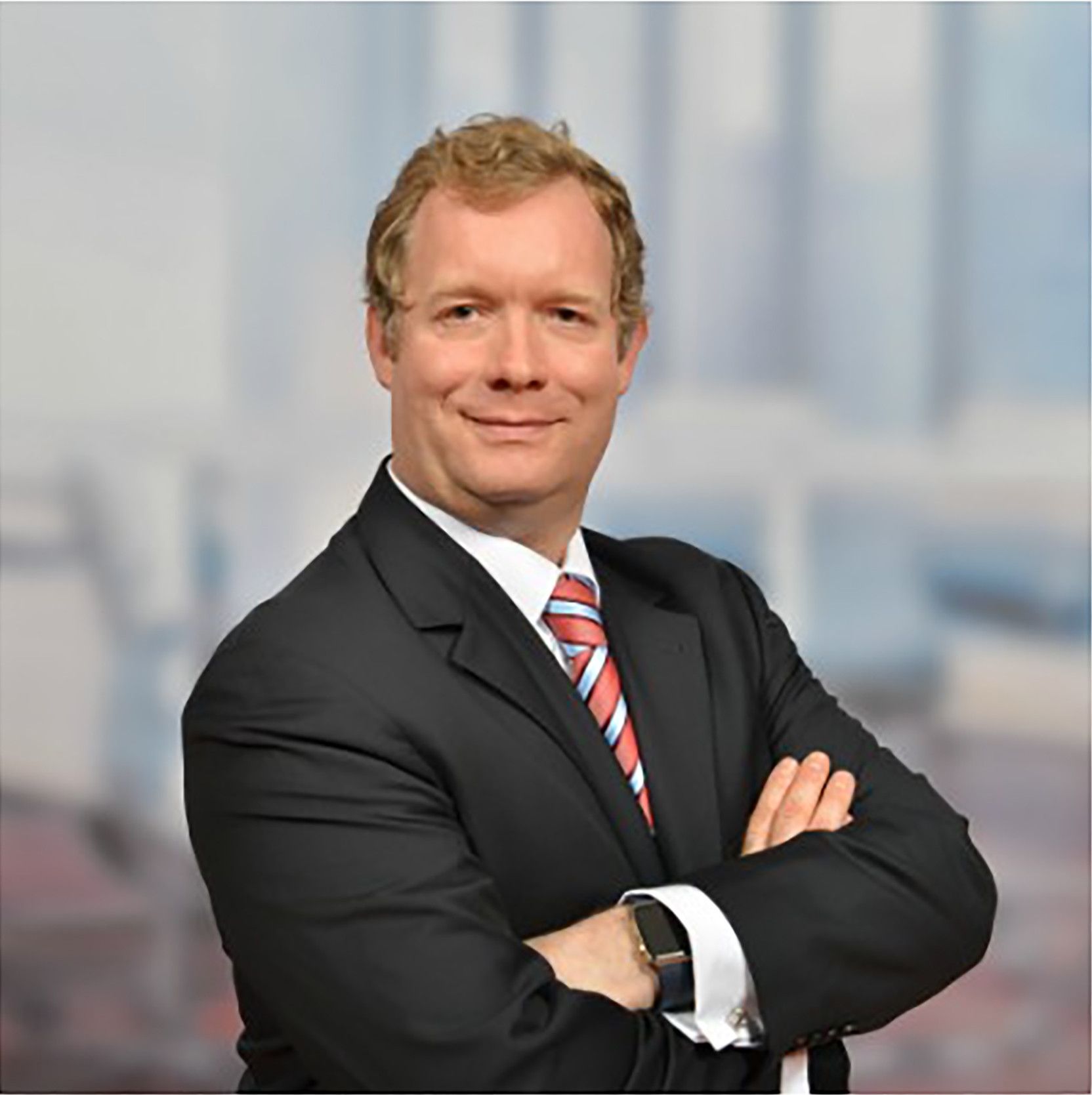 Christophe Lenaerts in a dark suit and red striped tie, arms crossed, against a blurred city backdrop