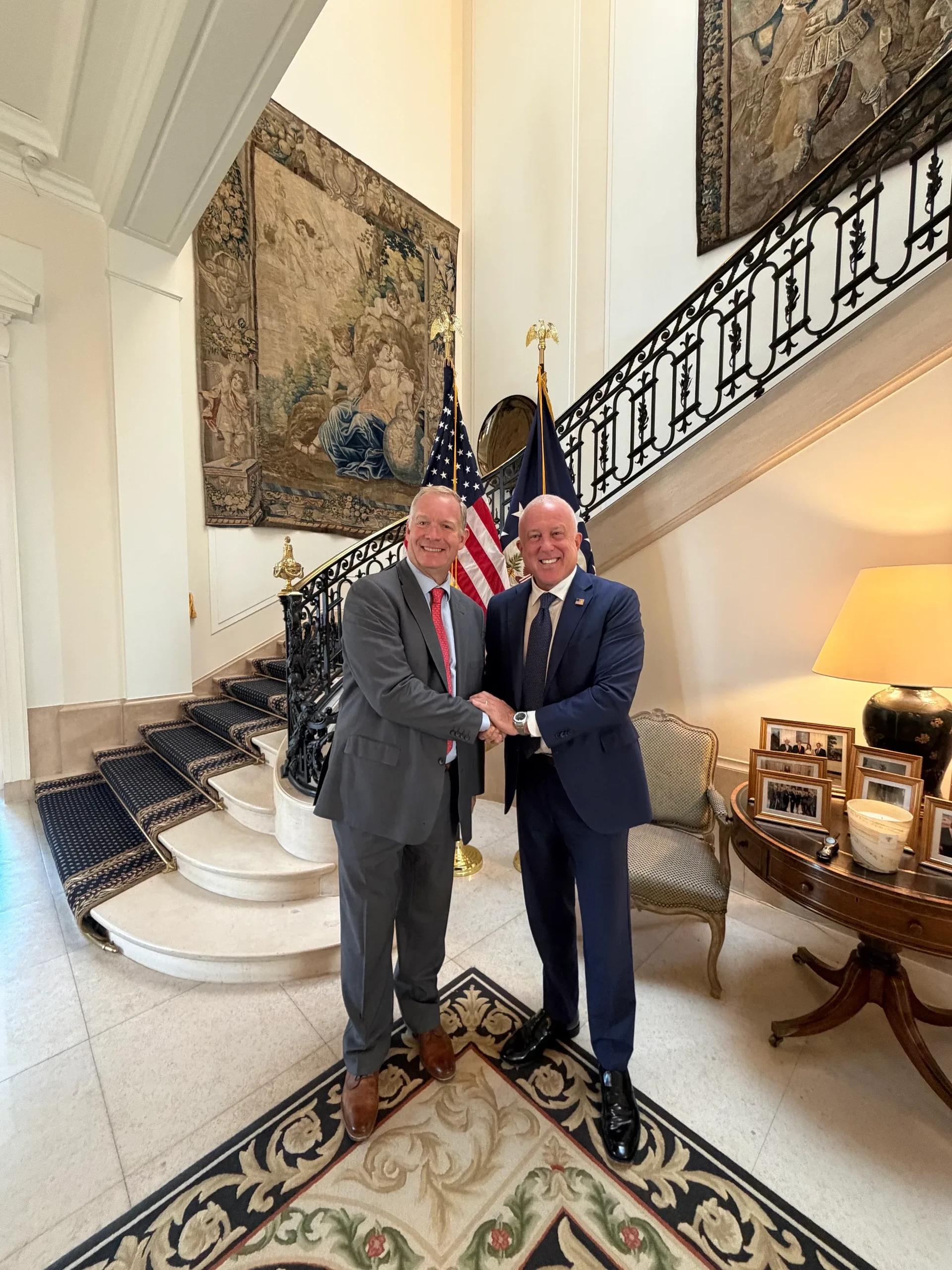 US Ambassador Bill White and Christophe Lenaerts shaking hands before American flags at a grand staircase