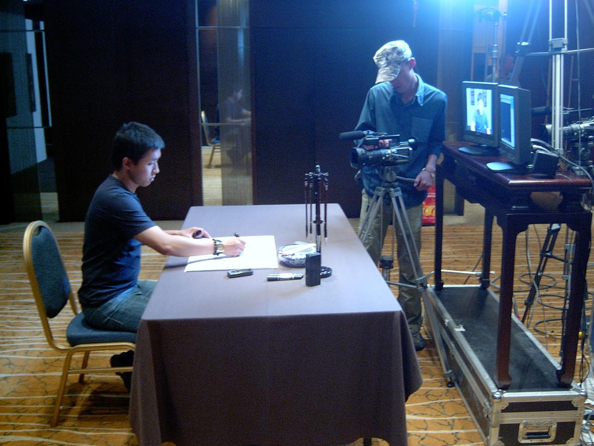 Man seated at a table with brushes and paper while a camera operator frames the shot beside two monitors